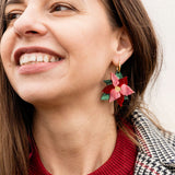 A woman smiling, wearing a red sweater and a plaid coat, shows off her festive jewelry—a poinsettia-shaped earring with red petals and green leaves, perfect as holiday earrings.