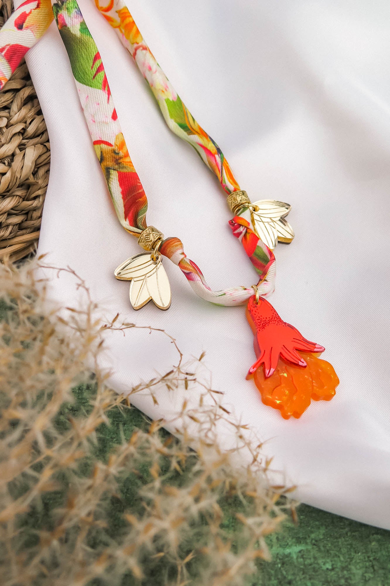 A colorful silk cord necklace with gold leaf charms and an orange-red hand-shaped pendant, displayed on white fabric with a woven basket and beige dried grass nearby.