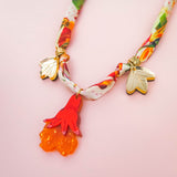 A colorful handmade necklace featuring two gold bee charms and a red-orange floral pendant, displayed on a soft pink background.