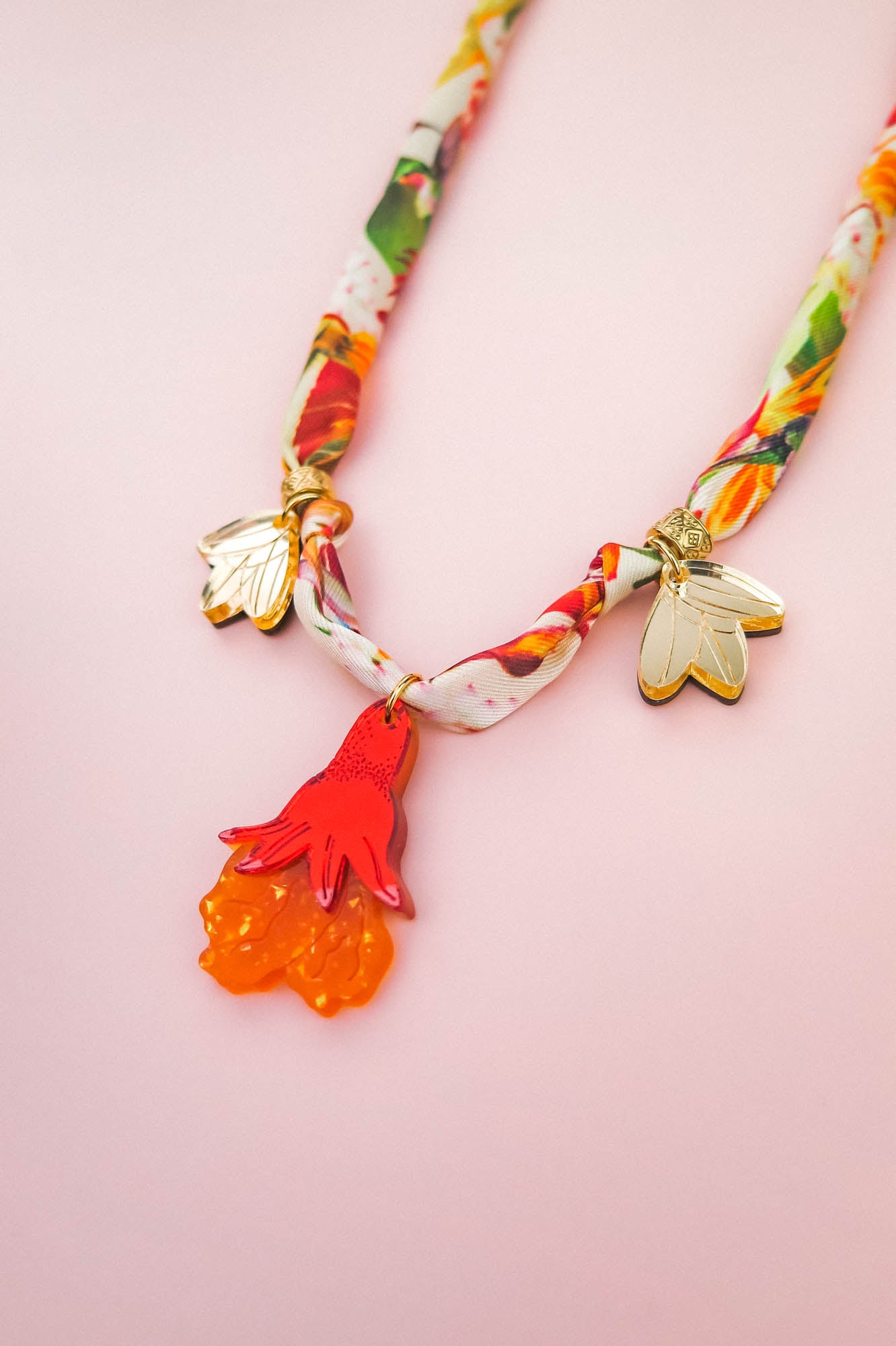 A colorful handmade necklace featuring two gold bee charms and a red-orange floral pendant, displayed on a soft pink background.