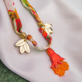 A colorful floral scarf with gold and white flower-shaped clips and a bright orange and pink pendant, displayed on a white fabric surface with a green background, pairs beautifully with a handmade pomegranate flower necklace.