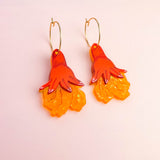 A pair of Little Turtle Hoop Earrings featuring gold hoops and pendants shaped like orange and yellow hand-drawn flowers, set against a pale pink background—beautifully handcrafted earrings for a playful, artistic touch.