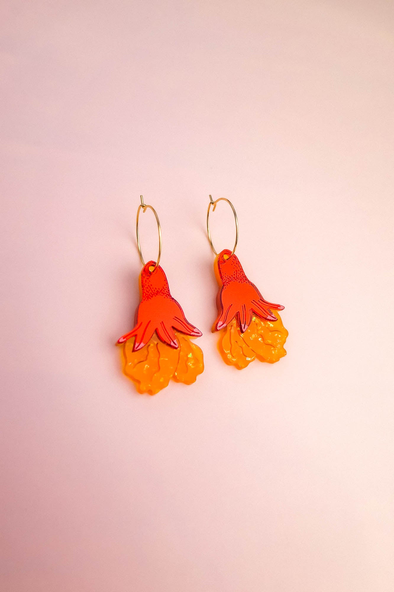 A pair of Little Turtle Hoop Earrings with gold hoops and dangling charms shaped like stylized orange and yellow flowers, displayed on a light pink background. These handcrafted earrings add a cheerful touch to any look.