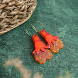 A pair of Little Turtle Hoop Earrings shaped like red hands with orange, ruffled details hang on gold hoops. The handcrafted earrings are placed on a green surface near a woven basket.