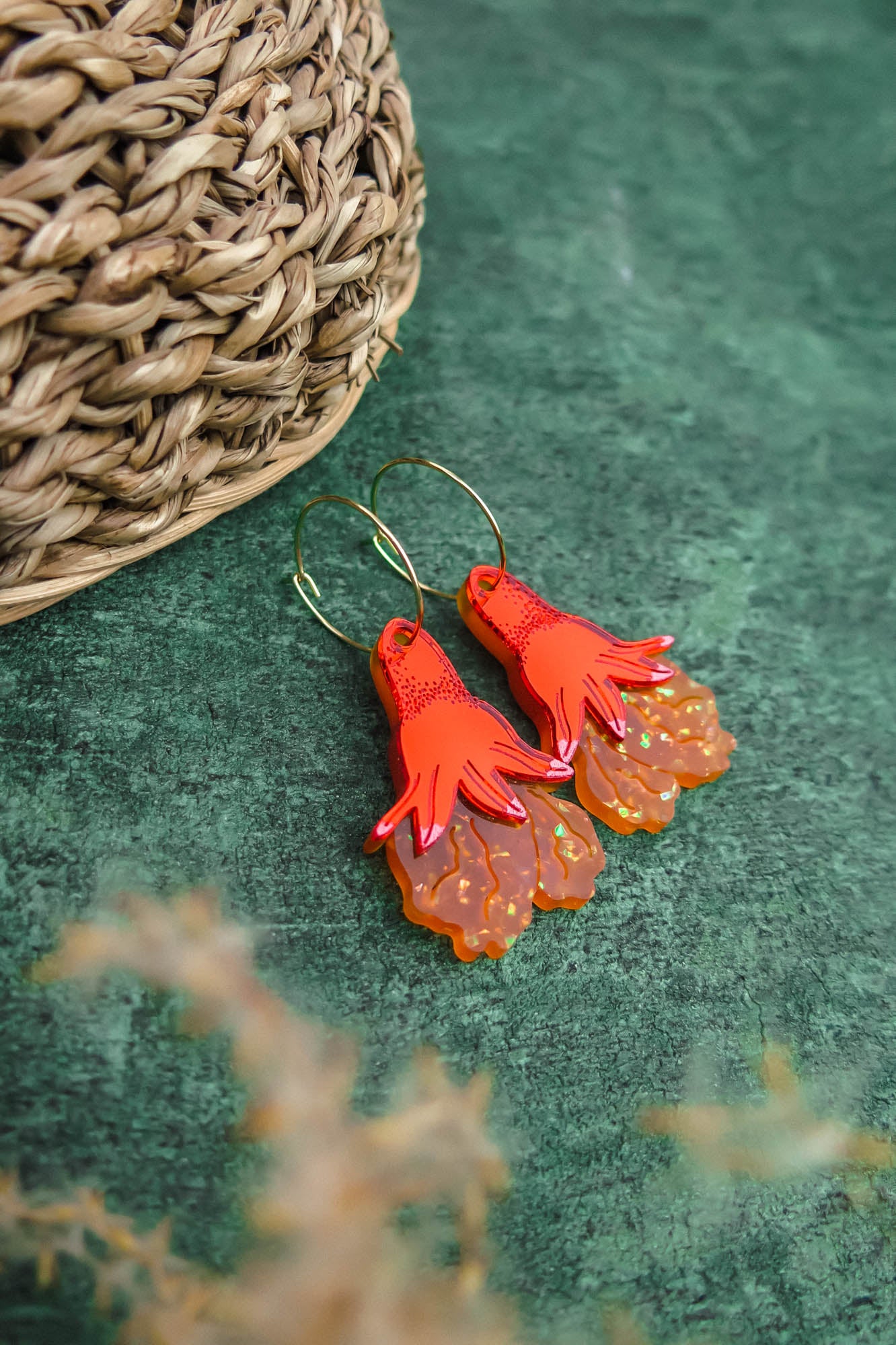 A pair of Little Turtle Hoop Earrings shaped like red hands with orange, ruffled details hang on gold hoops. The handcrafted earrings are placed on a green surface near a woven basket.