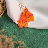 A pair of orange and yellow handcrafted earrings, shaped like stylized flowers or flames, rest on a white fabric backdrop. Green textured ground and part of a woven basket are visible, highlighting these unique gold hoops.