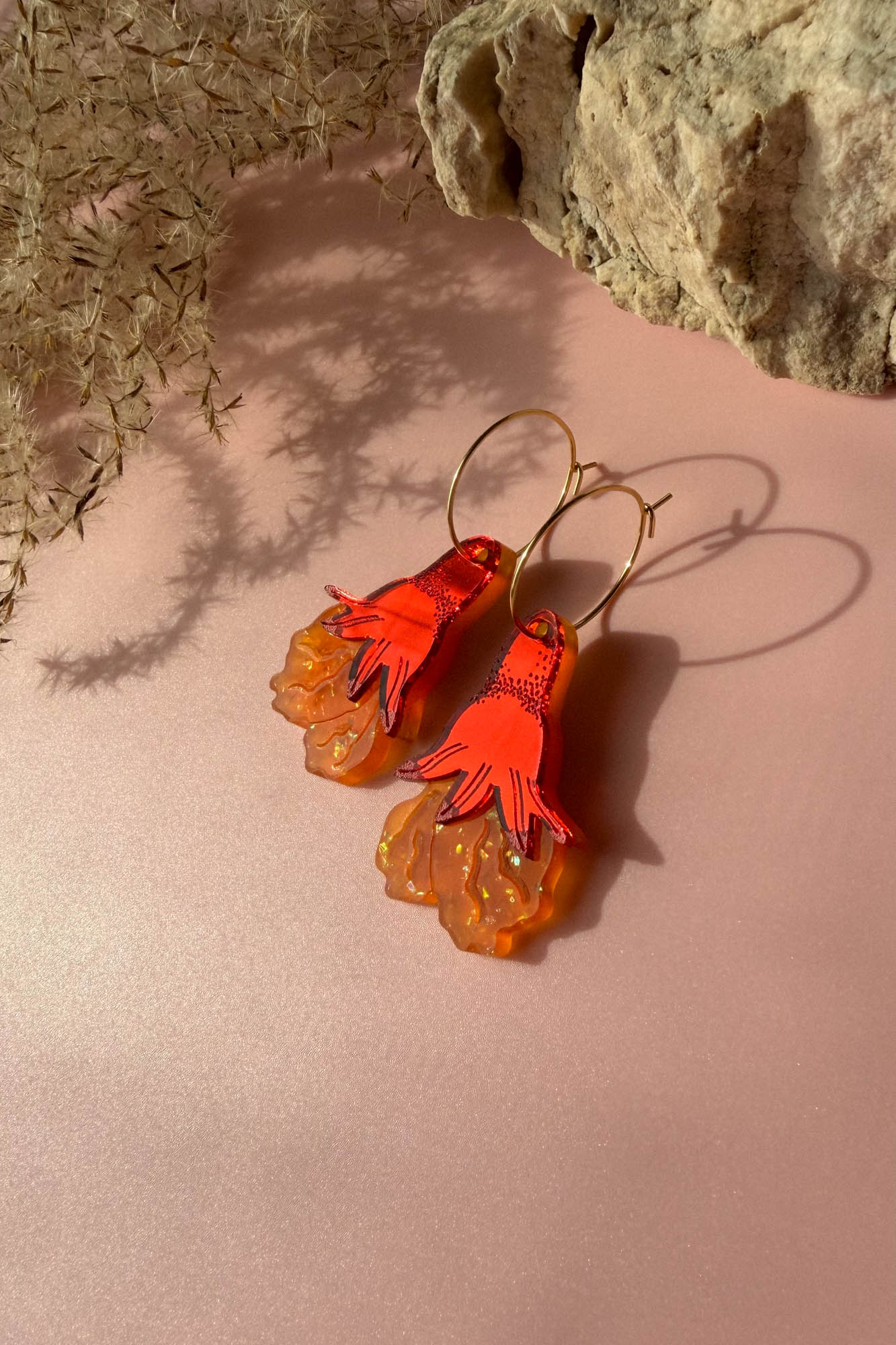 A pair of handcrafted earrings with orange and red hand-shaped charms and translucent, wavy orange accents dangle from gold hoops, resting on a pale pink surface near a rock and dried plant shadows.