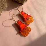 A pair of handcrafted earrings with orange hand-shaped charms and textured translucent orange bases dangle from gold hoops, displayed on a pink surface near a rock and dried plant.