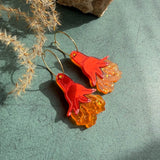 A pair of handcrafted earrings shaped like bright red-orange octopus tentacles with wavy, translucent lower sections, lying on a green surface near dried grass and a beige rock.