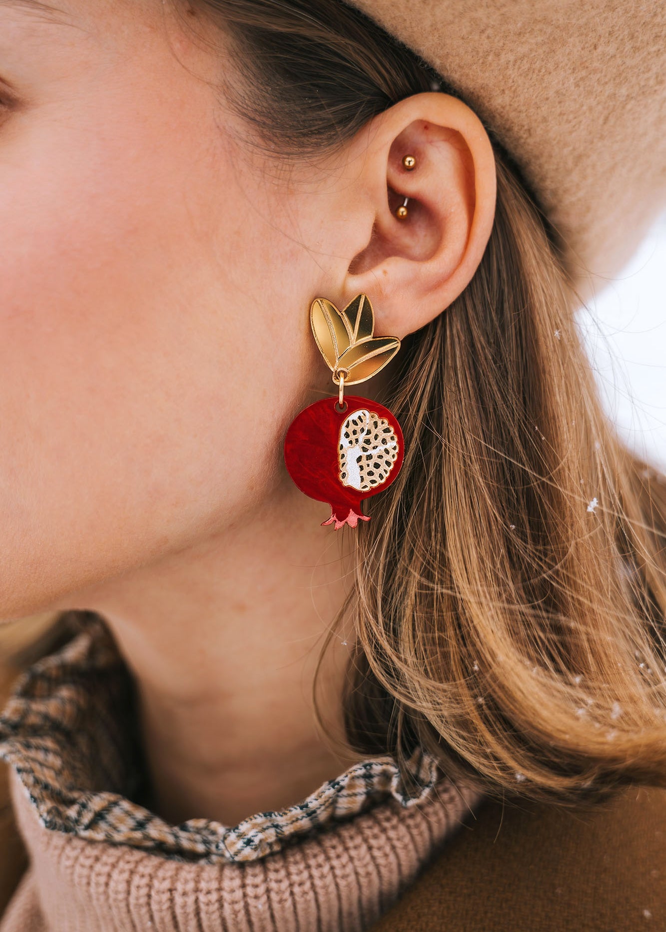 A close-up of a woman's ear wearing a gold stud and a large, handcrafted pomegranate-shaped earring with black leaves. She has a beige beret and a brown coat with a plaid collar. Snowflakes rest on her hair and coat, highlighting bold designs.