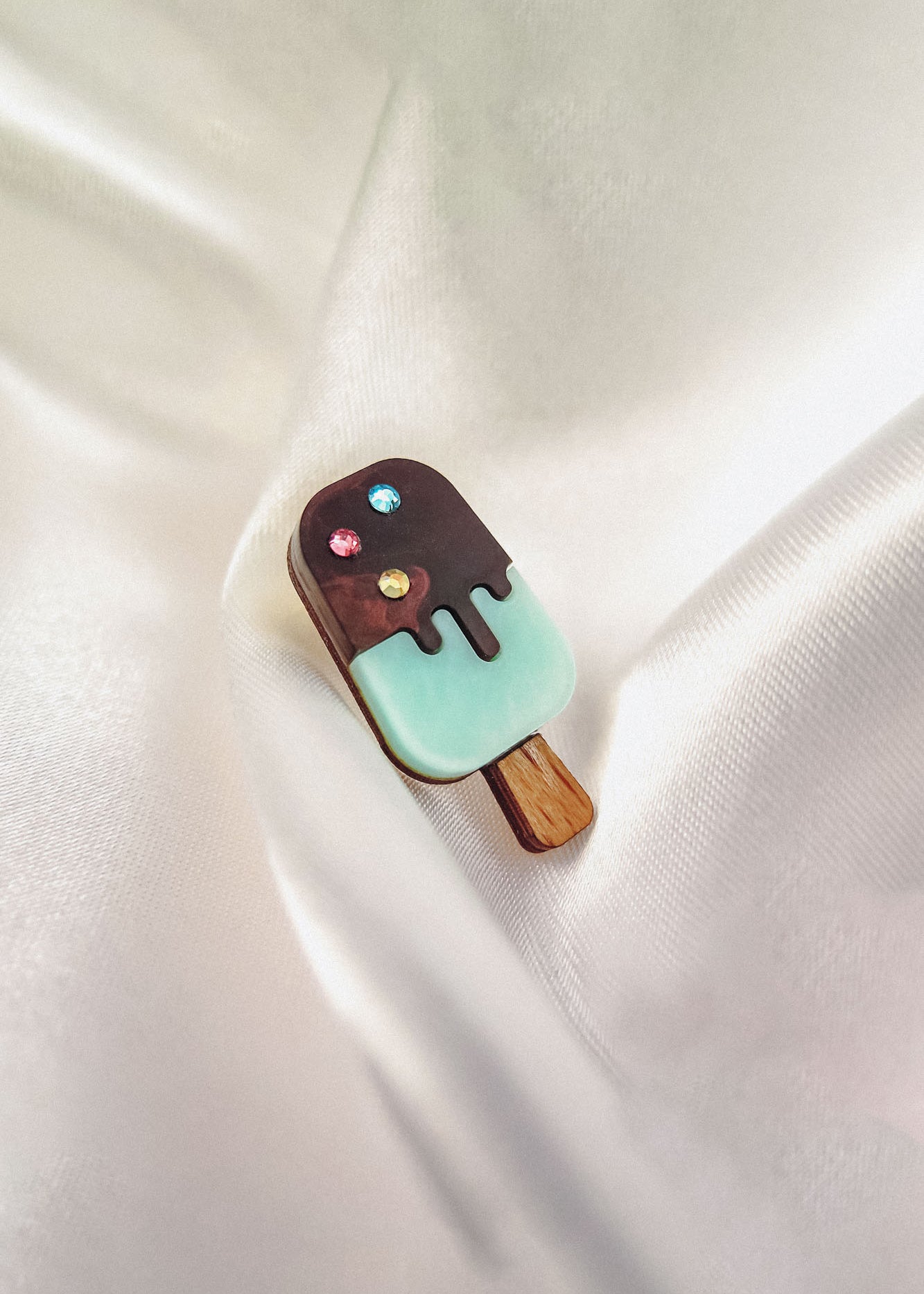 A handcrafted Popsicle Pin shaped like an ice cream bar with a wooden stick, chocolate coating, pastel blue bottom, and three colorful rhinestones sits atop smooth, white satin fabric.