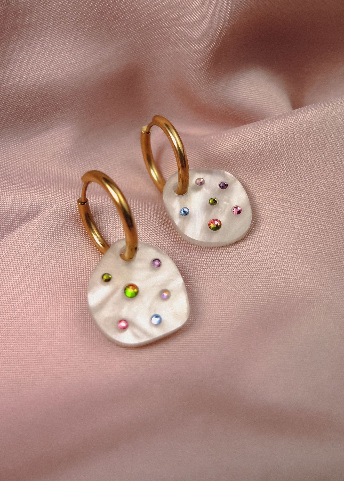 Lightweight hoop earrings with irregular white pearl-like drops, each decorated with colorful small gemstones, displayed on soft pink satin fabric.