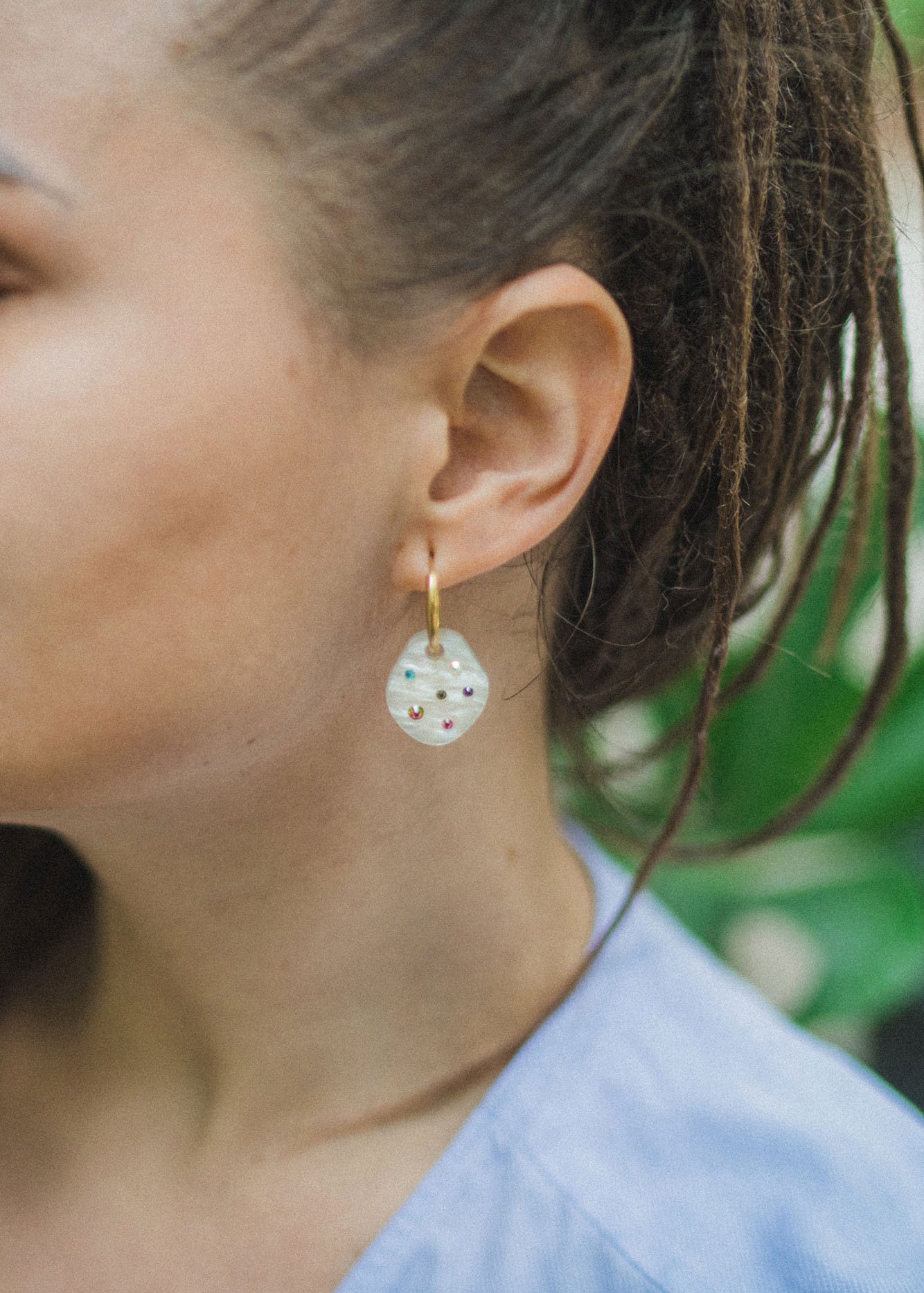 Close-up of a person with brown hair in dreadlocks wearing handcrafted, translucent round earrings with small colorful beads and a light blue top, set against a green leafy background.