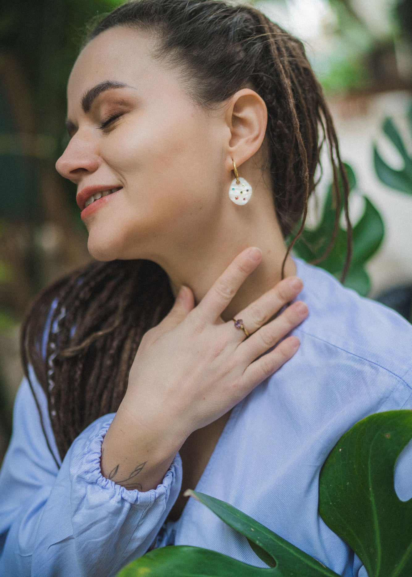 A woman with long braided hair wearing a light blue top smiles with her eyes closed, gently touching her neck and displaying a ring and handcrafted earrings, standing near green leaves.