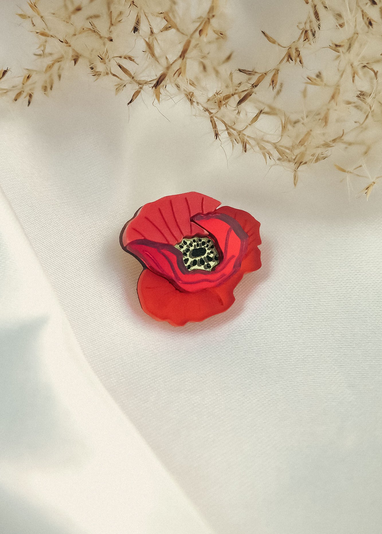 A handmade Red Poppy Flower Lapel Pin rests on a white fabric surface, with delicate dried grass or flowers in the background—an elegant accessory for fashion enthusiasts.