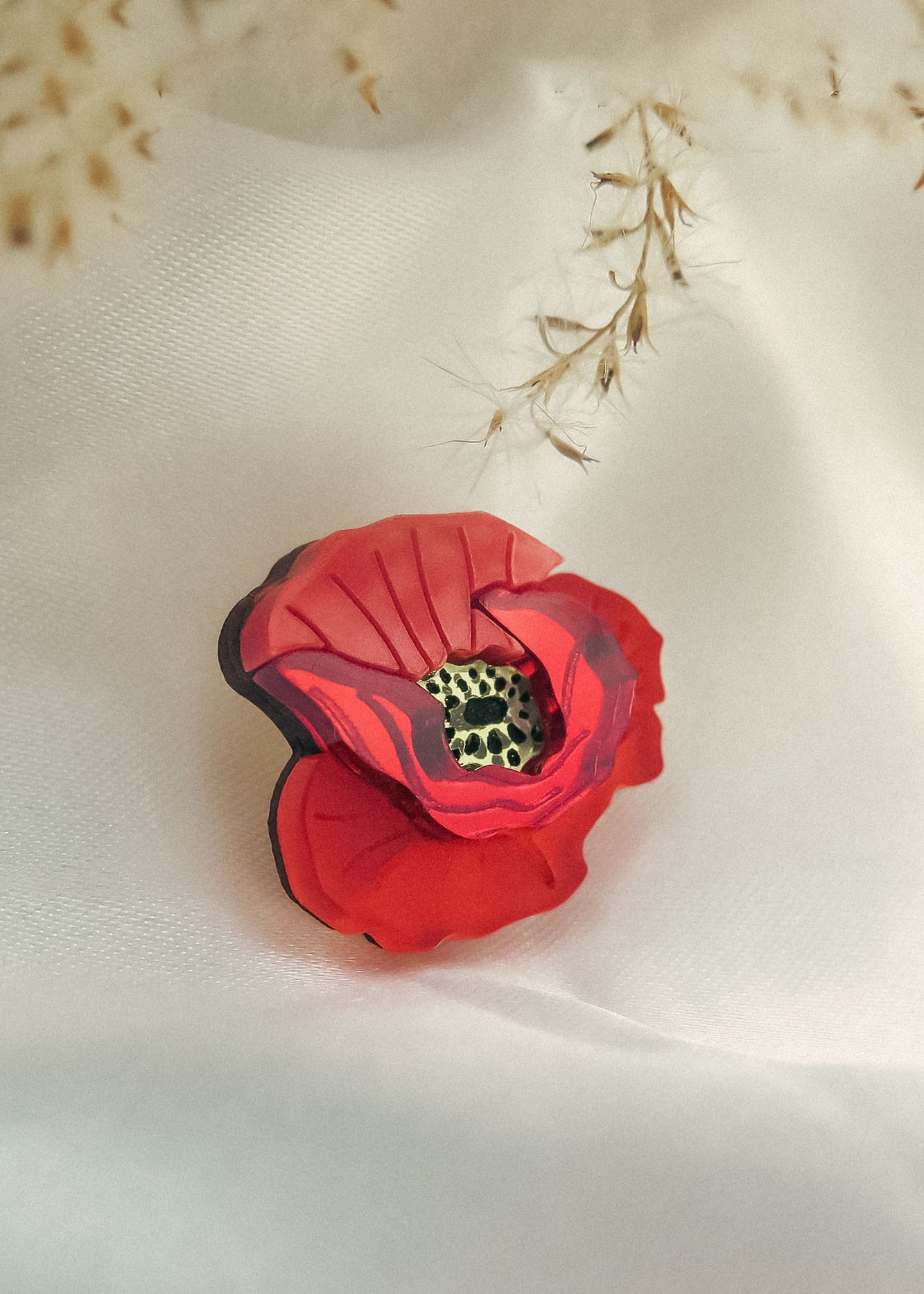 A handmade Red Poppy Flower Lapel Pin with a detailed textured center rests on soft white fabric, while delicate dried grass hangs above—an elegant accessory perfect for fashion enthusiasts.