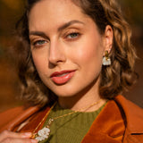A woman with wavy brown hair wears an elegant green top, brown jacket, and handcrafted gold jewelry with matching white flower-shaped earrings and a Tulip Necklace, holding the necklace and looking confidently at the camera.