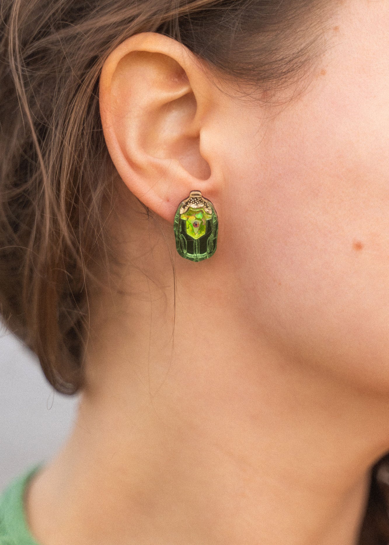 Close-up of a person wearing Rose Chafer Studs—nature-inspired earrings with green gemstones and gold details. The person has wavy brown hair, a green top, and is set against a softly blurred background.