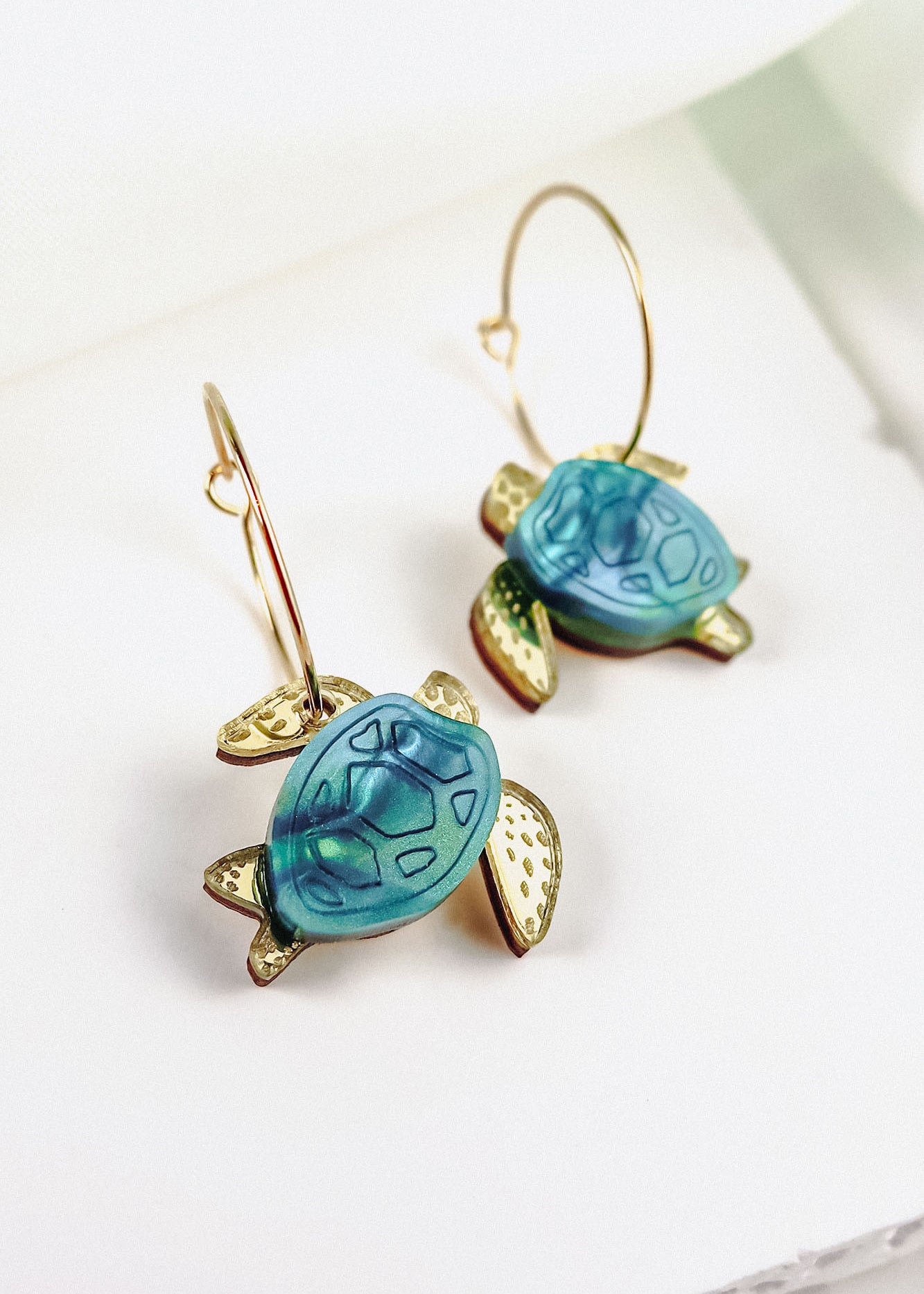 Gold hoops featuring blue and gold handcrafted turtle-shaped charms with detailed shells and flippers, the Little Turtle Hoop Earrings are displayed on a white background.