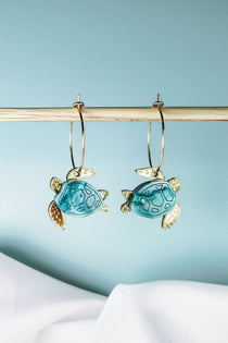 Two handcrafted gold hoops with hanging blue and gold turtle-shaped charms are displayed on a wooden rod against a light blue background, complemented by white fabric at the bottom. Discover the charm of our Little Turtle Hoop Earrings.