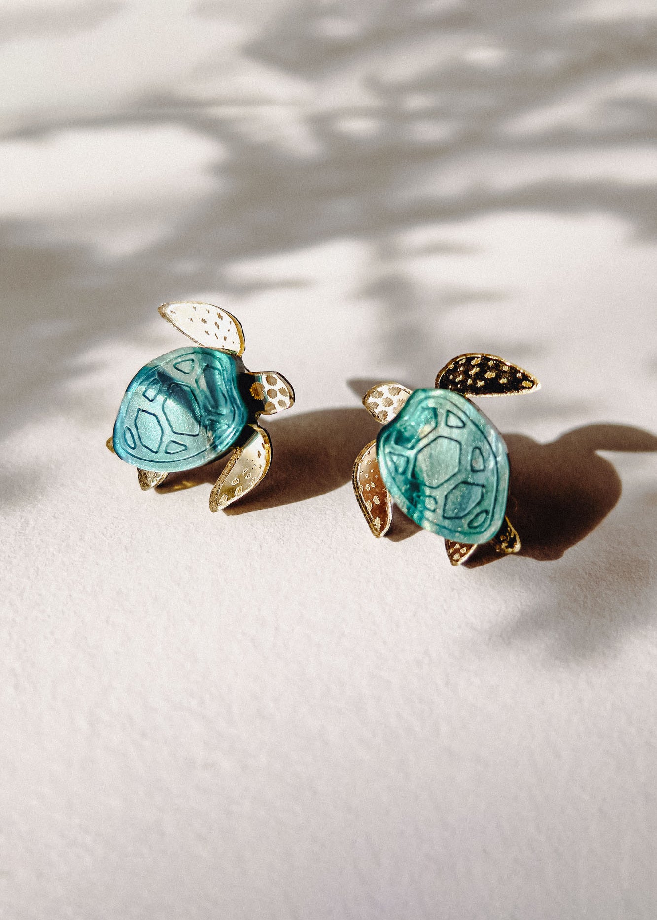 Two small Sea Turtle Studs with turquoise shells and gold-colored heads, legs, and tails rest on a light surface, casting soft shadows in natural light for a summery vibe.