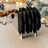 A small wooden sheep figurine made with black felt adds charm to your home decor, standing on a light wooden table with festive lights, pinecones, and greenery blurred in the background.