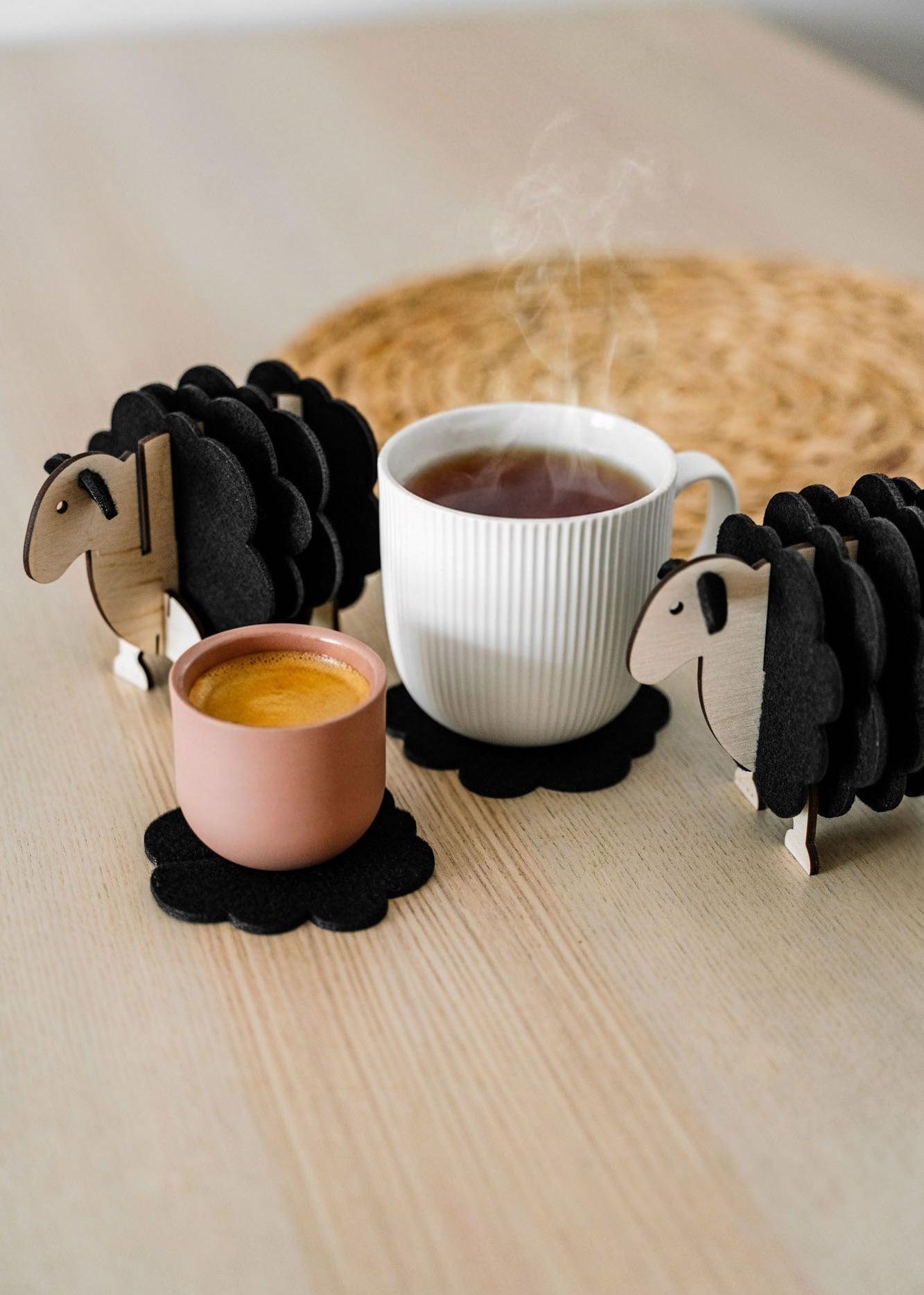 A white mug and a pink cup rest on felt coasters, accompanied by charming wooden sheep coaster holders on a light wooden table. Perfect as home decor accents, with a woven placemat in the background.
