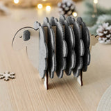 A wooden sheep-shaped coaster holder with felt coasters sits on a light wooden table, surrounded by pinecones, a snowflake decoration, and softly glowing fairy lights—charming home decor for cozy gatherings.
