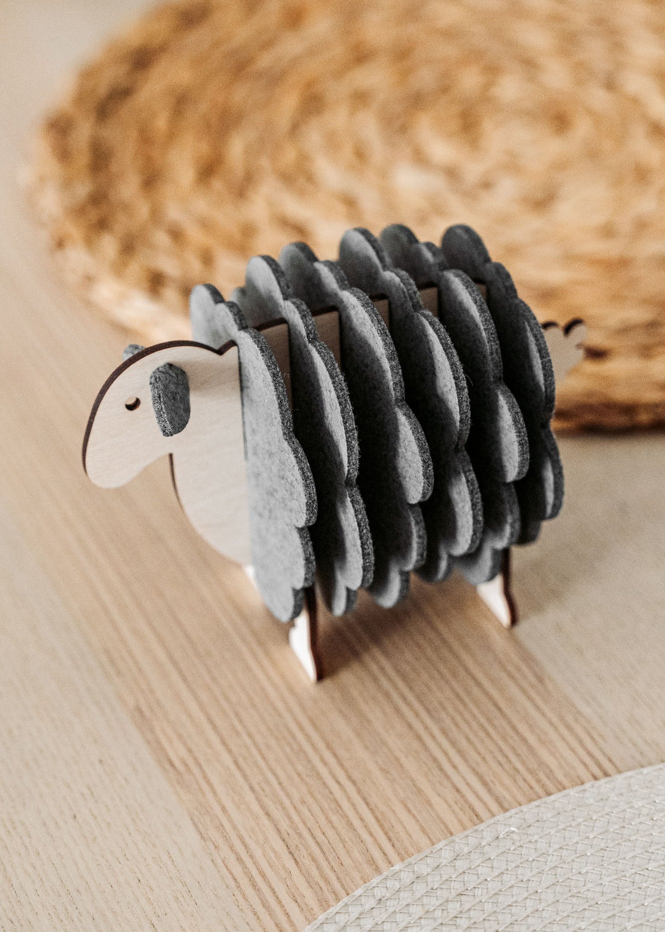 A small wooden sheep figurine with grey felt pieces for wool stands on a light wooden table, adding a charming touch of home decor, with a woven placemat in the background.