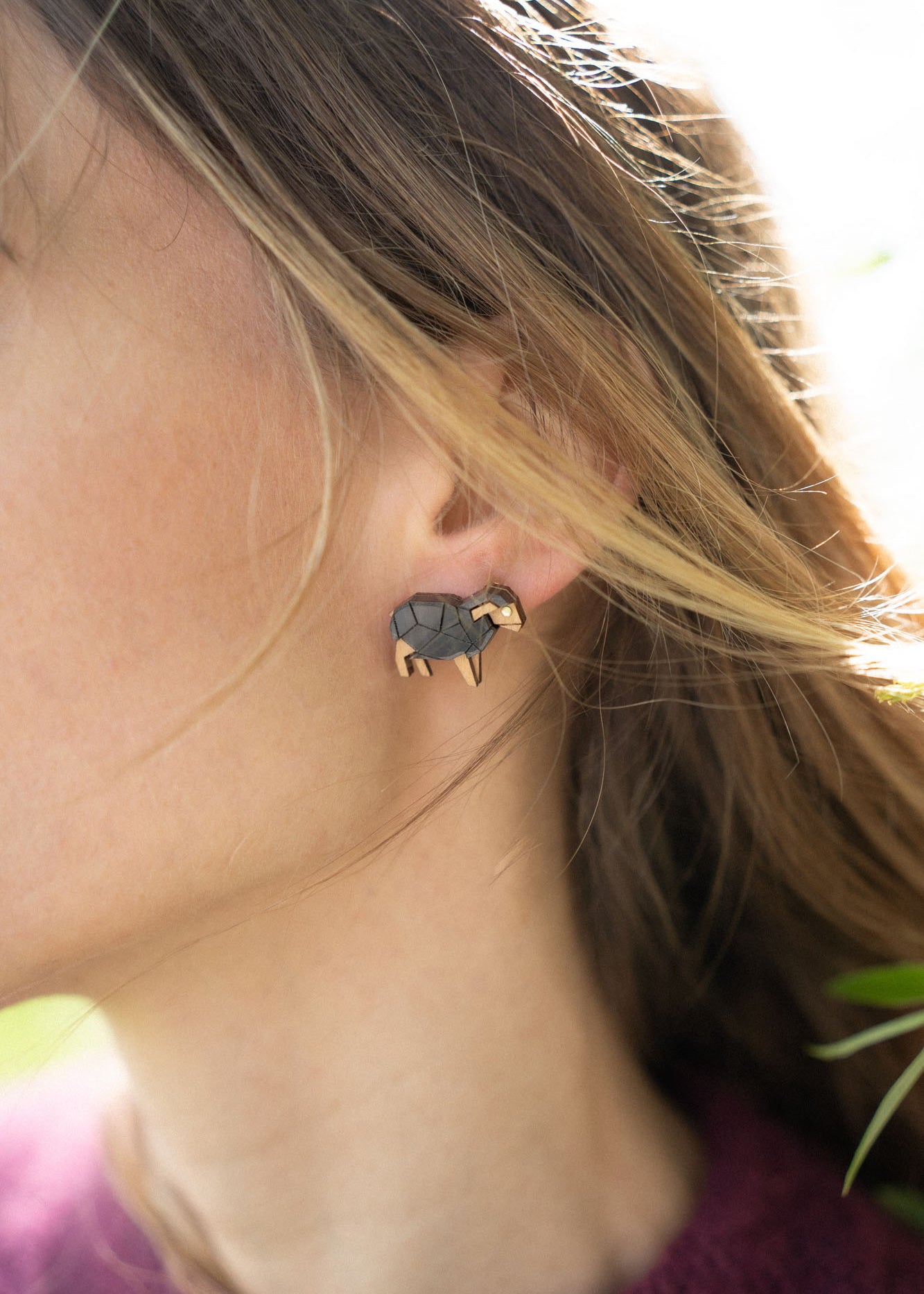 Close-up of a person wearing a small, black and gold elephant-shaped handcrafted earring adorned with sparkling rhinestones. Their brown hair partially covers the ear, and they are wearing a burgundy top against a softly blurred background.