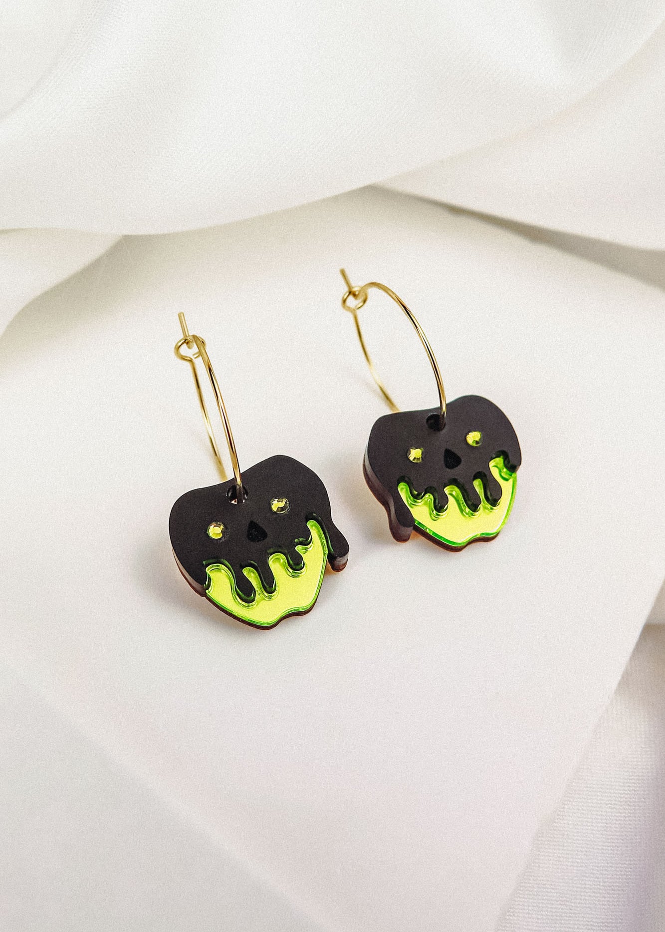 A pair of limited edition gold hoop earrings featuring black apple-shaped charms with bright green dripping accents, inspired by a Poisoned Apple Earrings Halloween design, displayed on a white fabric background.