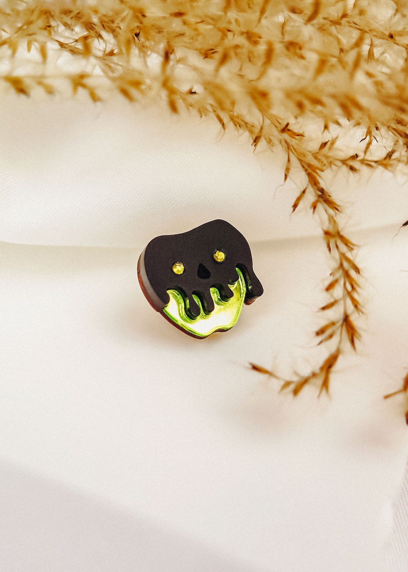 A small Poisoned Apple Badge shaped like a black skull with glowing green accents rests on white fabric, surrounded by delicate, dried plant stems—perfect for Halloween lovers and available in limited stock.