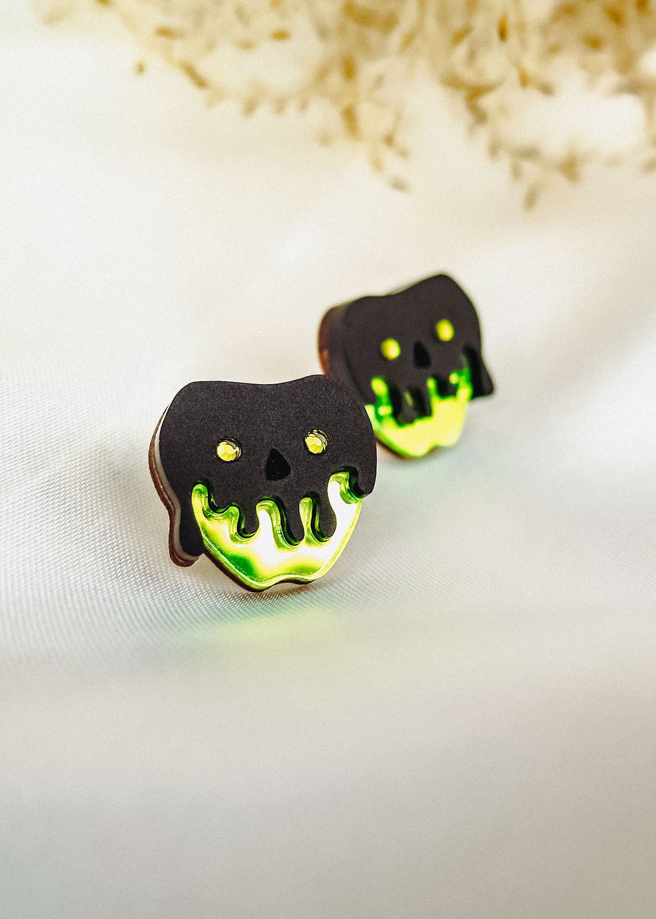 Two cookies decorated to look like poison apples, with black and neon green icing resembling a dripping skull, are placed on a light fabric surface. Their handcrafted details perfectly capture a Halloween look against the blurred floral background.