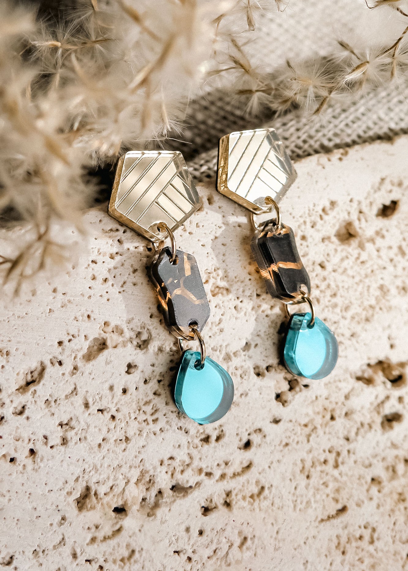 A pair of eco-friendly, handcrafted geometric dangle earrings with gold hexagon tops, marbled brown and black middle links, and translucent blue tear-drop earrings, displayed on a textured beige stone surface.