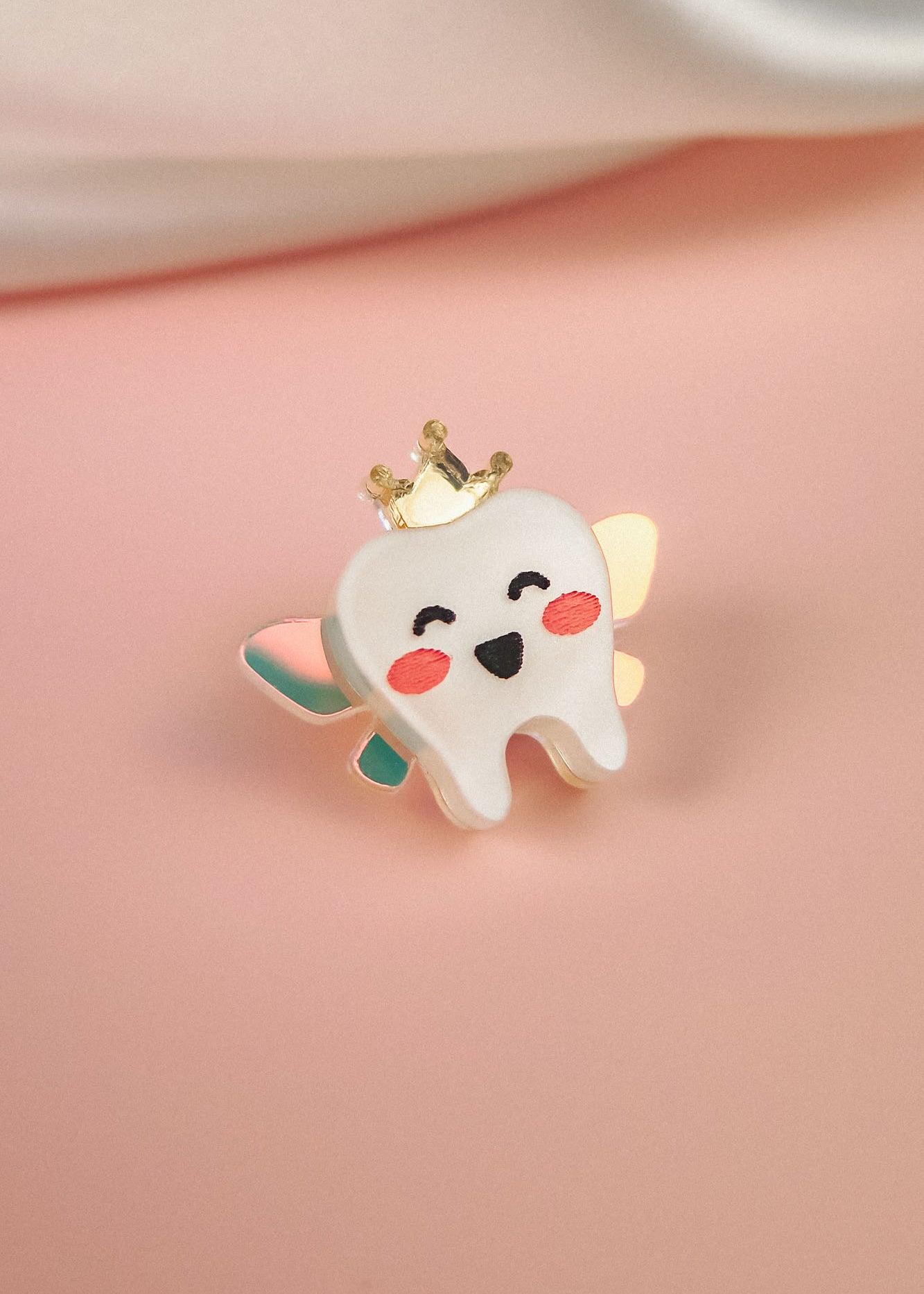 A cute Tooth Fairy Pin shaped like a smiling tooth with pink cheeks, a golden crown, and pastel wings celebrates childhood milestones, all handcrafted and set against a soft pink background.