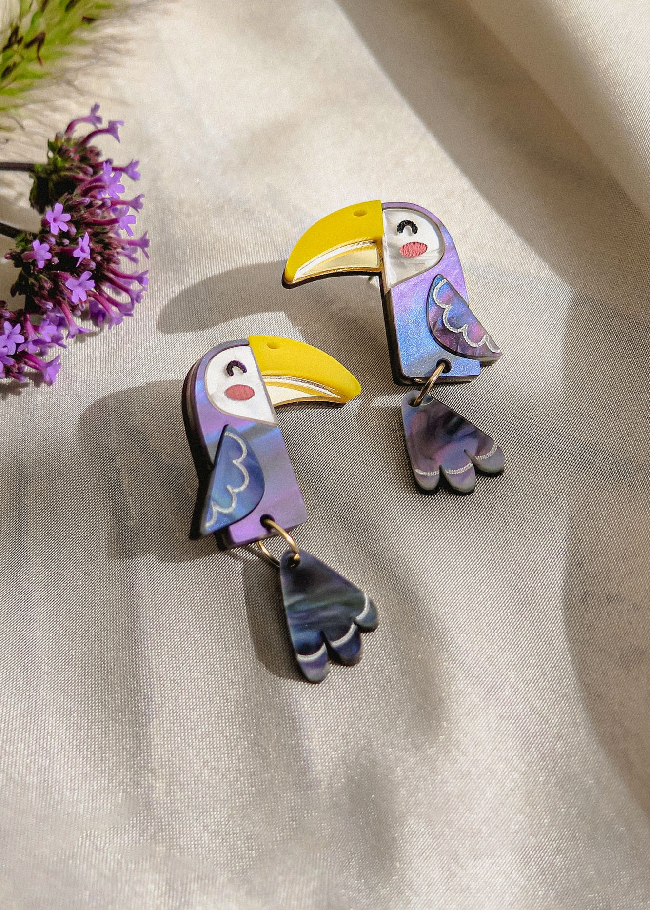 Two colorful Toucan Earrings made of iridescent materials rest on a light, satin-like fabric. Purple flowers are partially visible in the top left corner, adding to the playful pop of color in these handcrafted earrings.