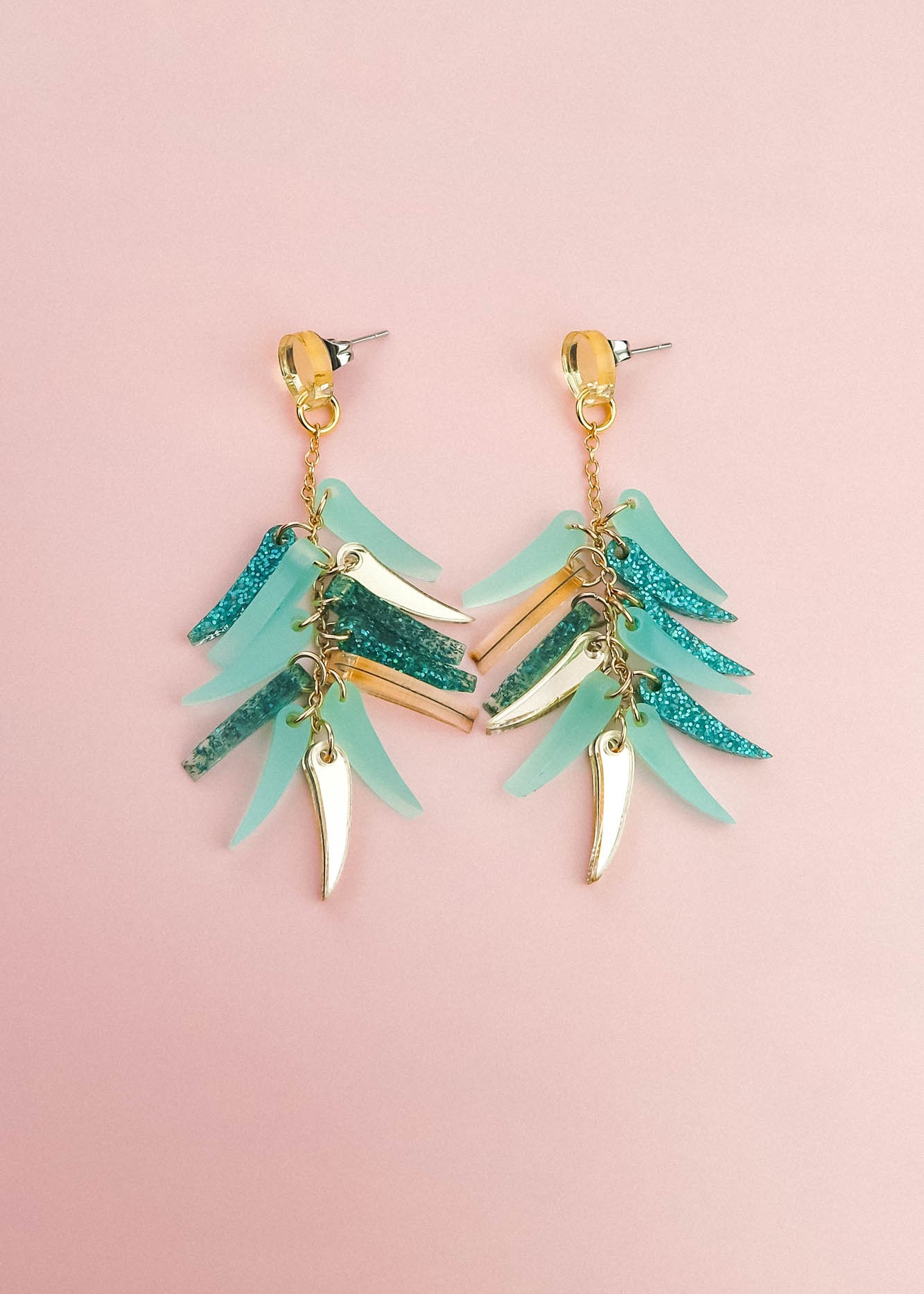 A pair of dangling earrings with gold hardware, featuring layered teal, blue glitter, and gold leaf-shaped charms, these unique style earrings are displayed on a light pink background.
