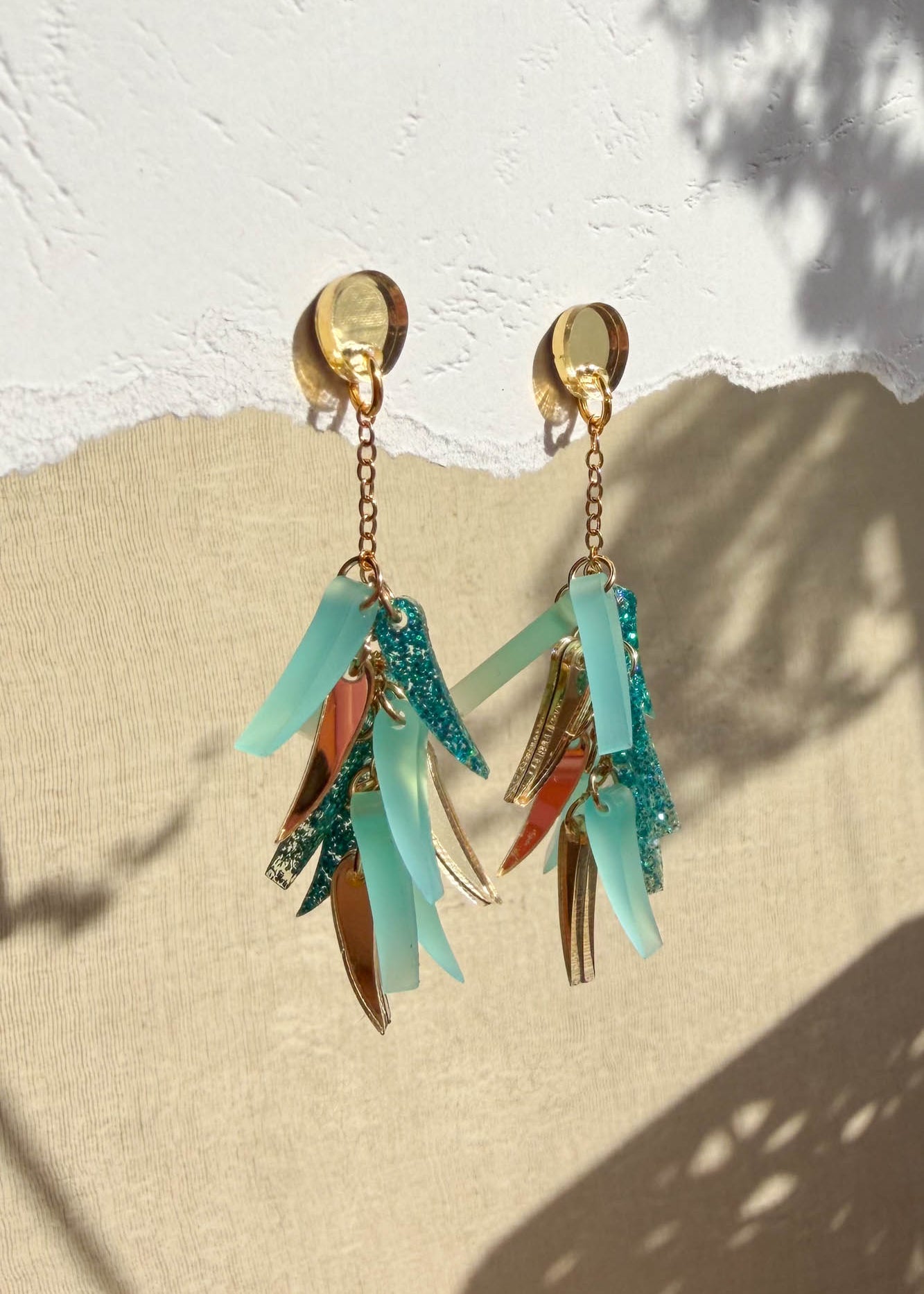 A pair of dangling acrylic earrings with gold, teal, and aqua-toned charms hang against a textured white and beige background, casting natural shadows that highlight their unique style.