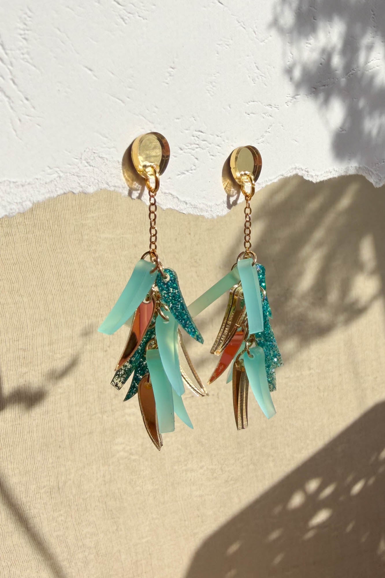 A pair of dangling acrylic earrings with gold, teal, and aqua-toned charms hang against a textured white and beige background, casting natural shadows that highlight their unique style.