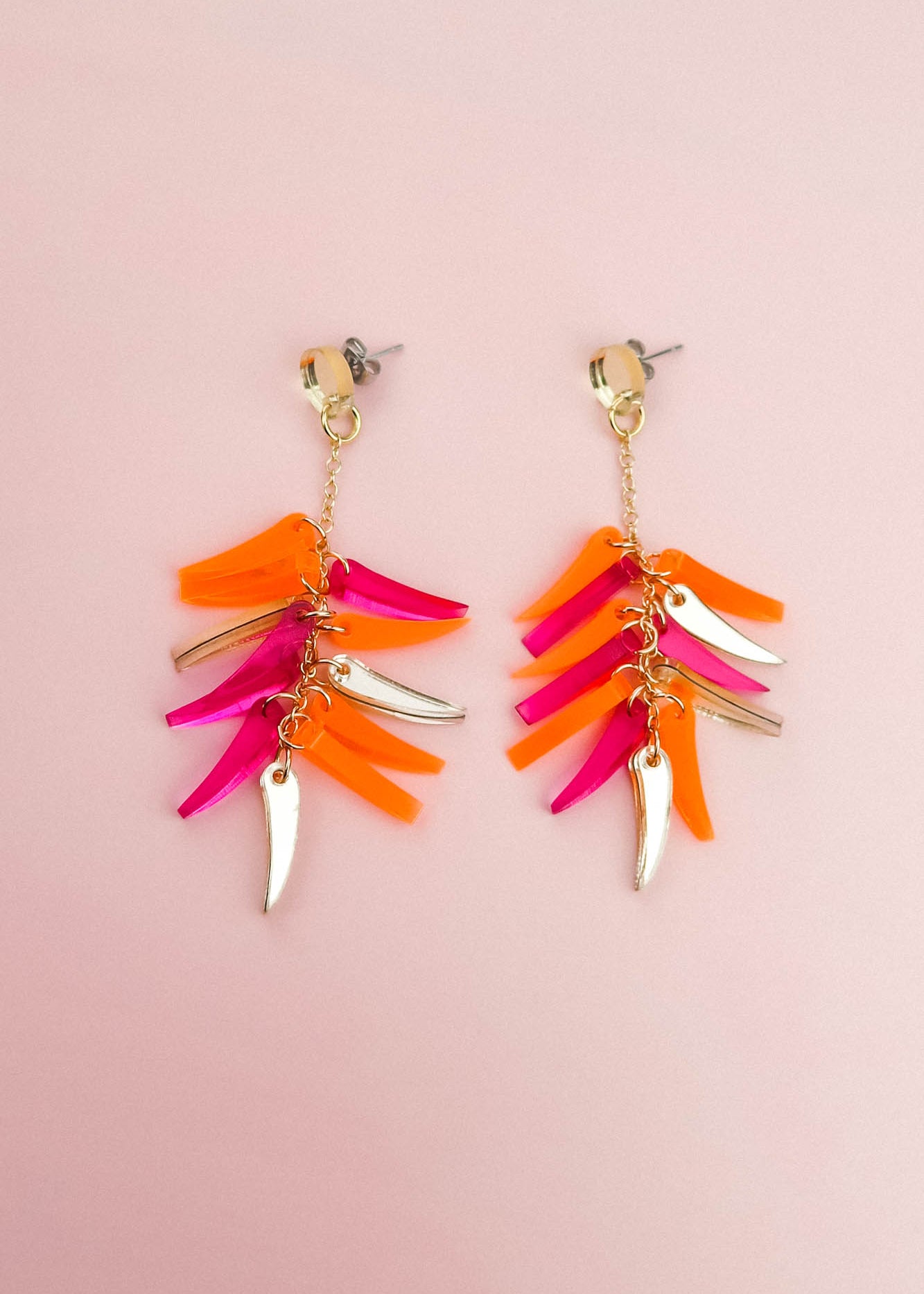 Gold dangle earrings with dangling pink, orange, and gold leaf-shaped charms arranged in layers, these featherlight earrings are displayed on a pale pink background.