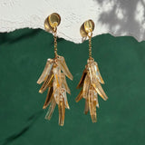 A pair of featherlight, gold chain dangle earrings with translucent, leaf-shaped pendants hang against a textured white and green background with shadow patterns.