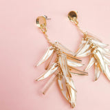 A pair of featherlight gold dangle earrings with leaf-shaped, translucent accents, displayed on a soft pink background.