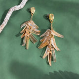 A pair of gold and clear acrylic dangle earrings shaped like layered leaves, these featherlight earrings are displayed on a green surface with textured driftwood and a partial glass dish.