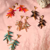 Four pairs of colorful, featherlight dangle earrings in orange, gold, turquoise, and black are arranged in a circle on a pink surface with a rock and dried grass nearby. Sunlight casts soft shadows across the scene.