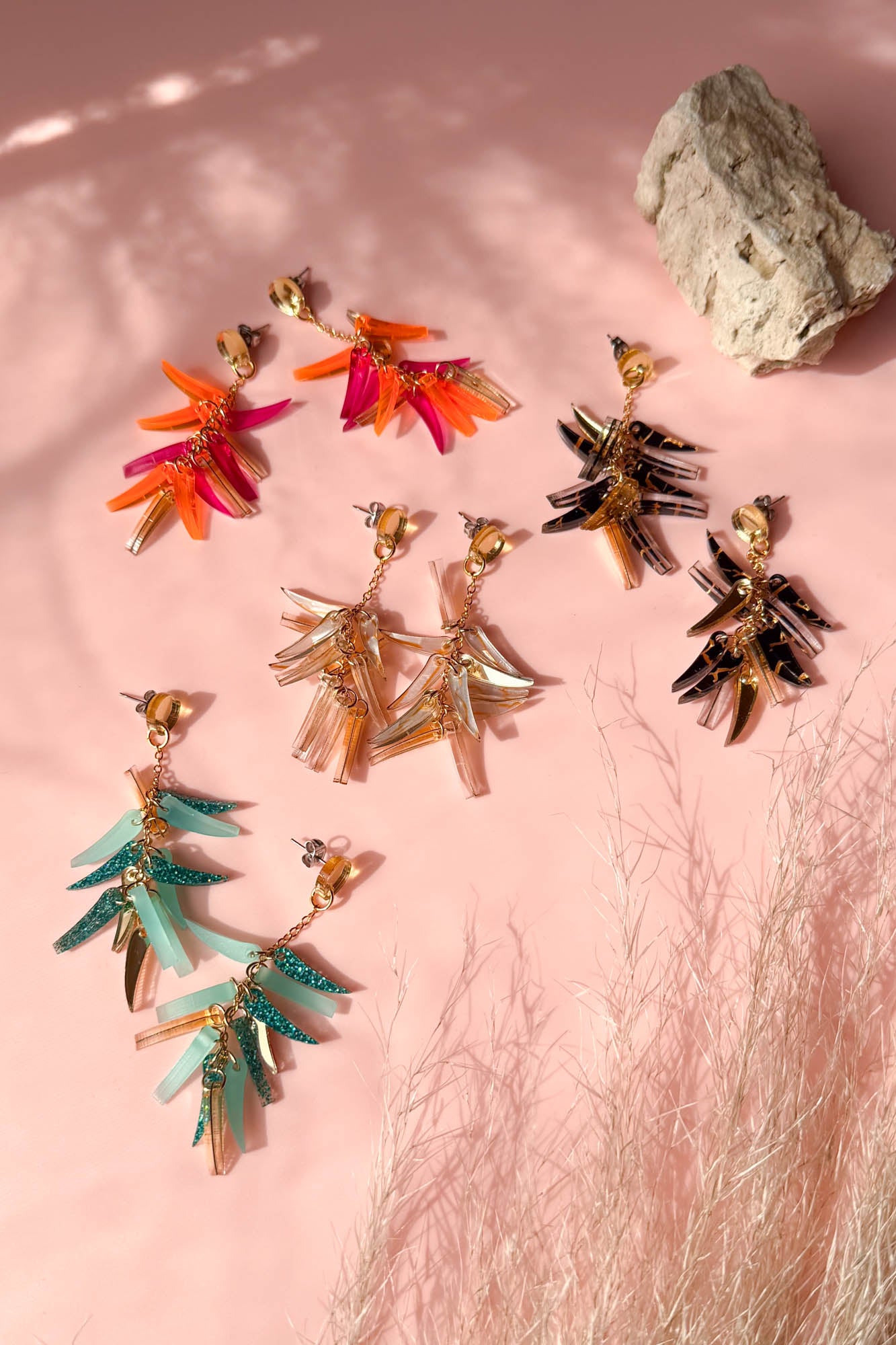 Four pairs of colorful, featherlight dangle earrings in orange, gold, turquoise, and black are arranged in a circle on a pink surface with a rock and dried grass nearby. Sunlight casts soft shadows across the scene.