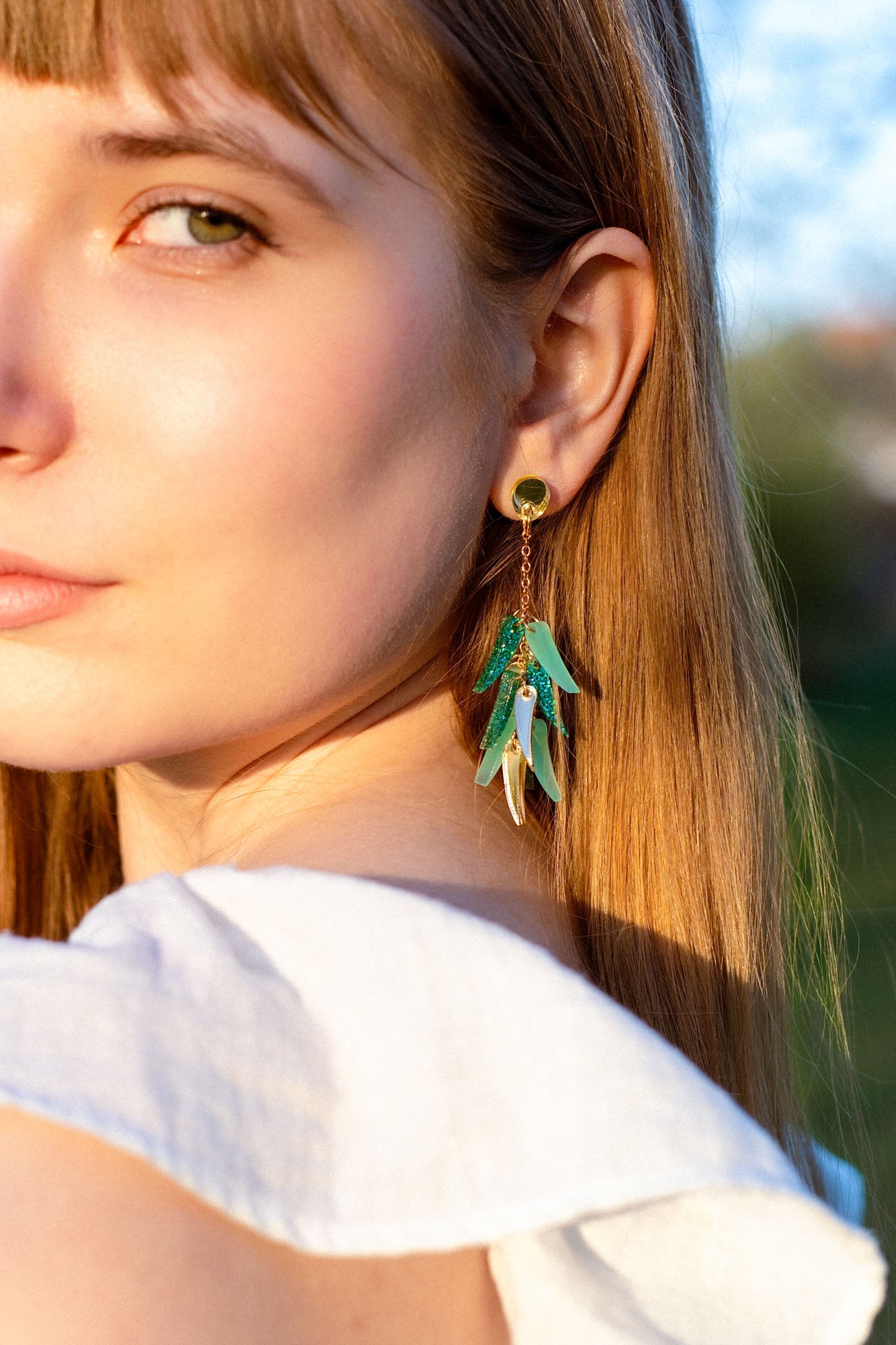 Close-up of a young woman with long light brown hair in a white top, showcasing featherlight dangle earrings shaped like green and gold leaves. She gazes slightly to the side, bathed in natural sunlight.