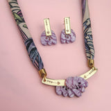 A patterned fabric handmade necklace with gold accents and a pendant featuring marbled purple shapes, paired with matching marbled purple earrings, is displayed on a soft pink background.