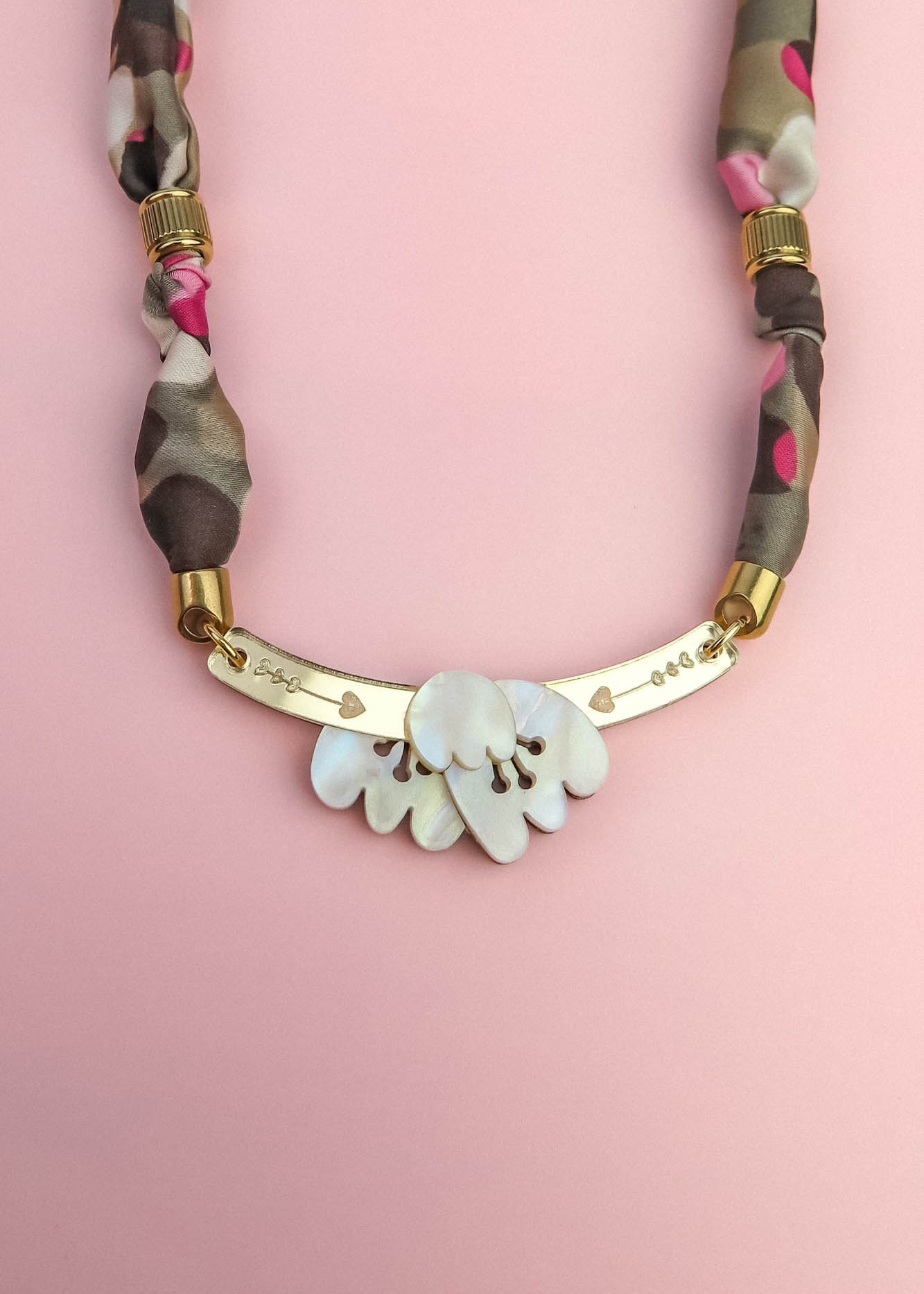 A handmade necklace with a gold curved bar, white shell-like pieces, and a multicolored fabric chain reminiscent of a silk-print scarf in green, pink, brown, and gold accents, displayed on a light pink background.