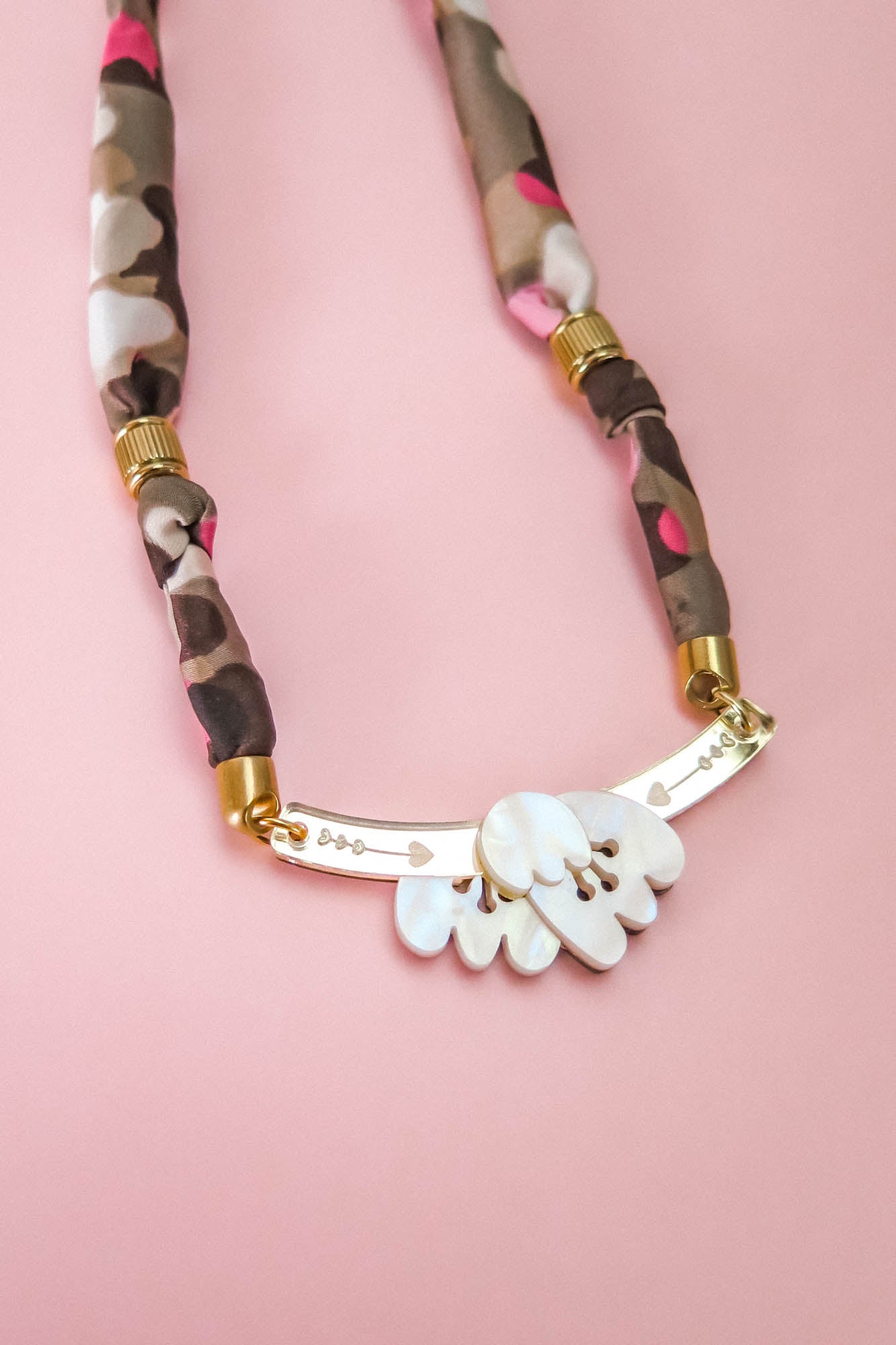 A handmade necklace with a pink, brown, and beige fabric band, gold accents, and a curved gold bar adorned with five white shell-like charms is displayed on a pink background.