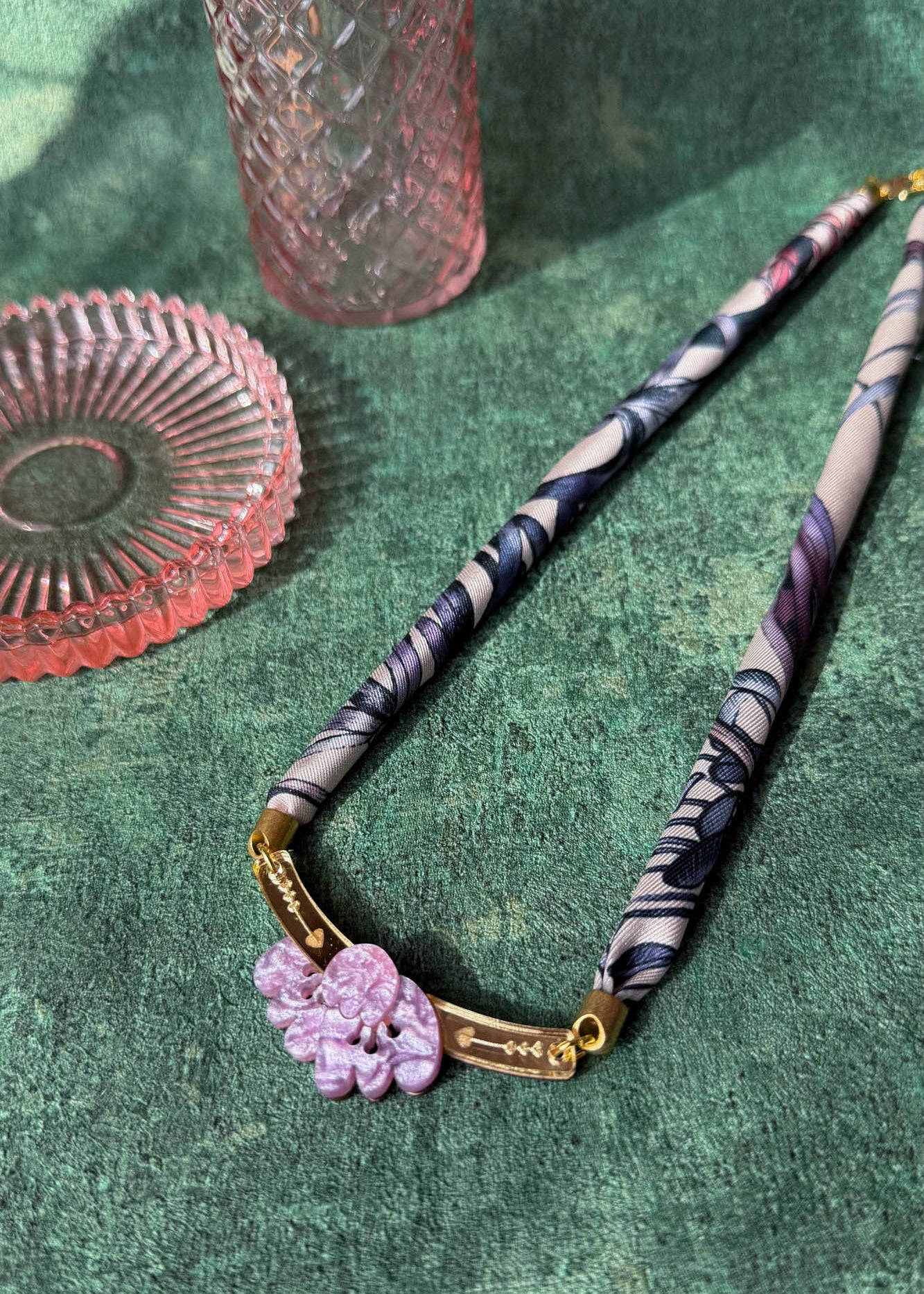 A handmade necklace featuring a patterned fabric and pink carved pendant with gold accents is displayed on a textured green surface, beside two pink glass dishes.